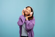 Excited asian woman listening to music with closed eyes using wireless headphones, standing over blue background