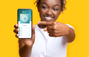Cheerful Young Black Lady Pointing At Smartphone With Opened Music App Interface