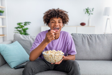 Canvas Print - Cheery African American teenager with bowl of popcorn enjoying funny movie, laughing at home