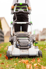 Caucasian Woman Wearing Boots And Overalls Is Mowing The Lawn In Late Fall. She Is Operating An Electrical Walk Behind Device. She Goes Over Fallen Leaves To Clear The Yard.