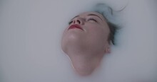 Beautiful Fashion Model Girl In Milk Bath, Touching Face Skin, Spa And Skin Care Concept. Beauty Young Woman With Red Rose Flower Petals Relaxing In Milk Bath, Rejuvenation. Slow Motion UHD Video.