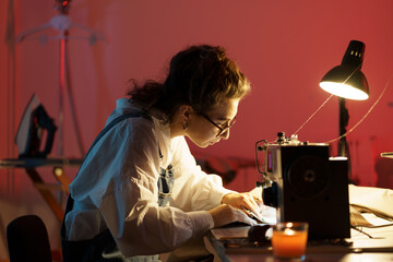 Wall Mural - Fashion designer sew clothing. Young woman busy working on sewing machine in studio. Self-employed tailor preparing for new clothes collection presentation in workshop showroom. Small business concept