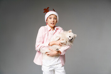 Wall Mural - Cute teenage boy in hat with antlers plays with his spitz dog on light background. New year and christmas time concept. Emotional facial expression. Caucasian guy happy on vacation