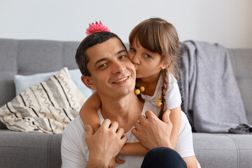 Indoor shot of happy man and little daughter hugging father with having funny hairstyle, looking camera, charming girl kissing dad's cheek, family expressing love and positive emotions.