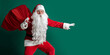 Emotional Santa Claus sneaking while carrying huge red sack with presents on green studio background