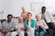 Smiling nurse sitting near interracial pensioners with yarn and digital tablet in nursing home