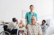 © LIGHTFIELD STUDIOS - Smiling nurse looking at camera near senior patient and interracial people in nursing home