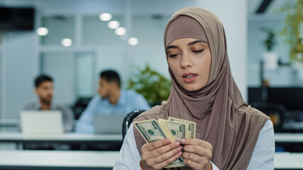 Serious concentrated eastern muslim girl in hijab office worker manager or accountant holding bundle of dollars in hands carefully counting cash paper money on payday keeps records of company income