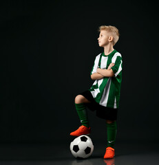 Wall Mural - Young soccer player, football school student, kid boy in red white striped uniform stands holding arms crossed and with ball under his foot and looks aside at copy space over black background