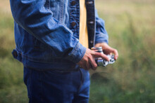Male Hands Holding Retro Photo Camera On Green Nature Background. Film Grain Effect. Man In Jeans