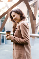 Wall Mural - smiling woman in beige blazer holding coffee to go outdoors