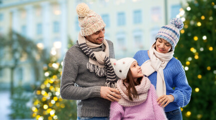 Poster - family, people and winter holidays concept - happy mother, father and little daughter in knitted hats and scarves over christmas tree lights on city street background