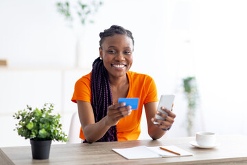 Young African American woman buying online with credit card and mobile phone, sitting at desk indoors