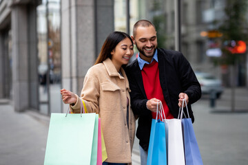 Wall Mural - Affectionate multinational couple standing near supermarket, looking inside shopper bags, going shopping together