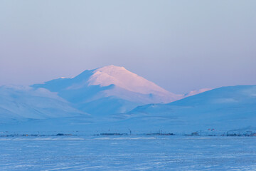 Wall Mural - Winter Arctic landscape. View of the snow-covered tundra and snow-capped mountains. The top of the mountain is illuminated by the setting sun. Northern settlement in the distance. Chukotka, Russia.