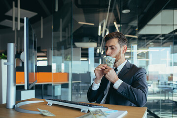 Joyful business man in sitting at desk working on laptop computer in bright office Achieving business career concept. Keep cash by making a gesture to the winner