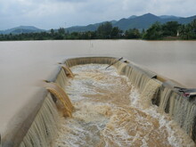 Brown Water In The Dam Overflowing Into The Spillway With Mountain And Green Forest In Background , Flood In Rainy Season, Thailand