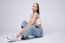 High Fashion Photo Of A Beautiful Elegant Young Woman In A Pretty White Top And Sneakers, Blue Denim Jeans Posing Over White, Soft Gray Background. Studio Shot. The Model Is Sitting