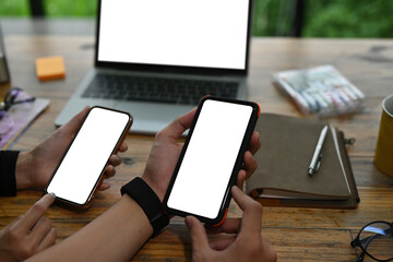 Poster - Close-up image of two hands holding a white blank screen smartphone over the working desk surrounded by an empty screen computer laptop and various stuff.
