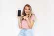 © Марина Демешко - A young caucasian amazed smiling blonde woman in a pink t-shirt holds mobile phone in her hand and points at black screen demonstrating copy space for text or design isolated on a white background