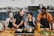 © Seventyfour - Portrait of female chef frying meat during cooking class with diverse group of people in kitchen interior