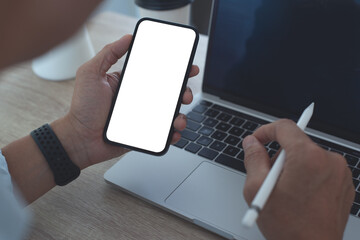 Canvas Print - Mockup image of mobile phone, business man hand holding blank screen mobile phone during working on laptop computer at home office