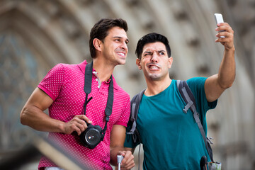 cheerful men tourists with suitcases are making selfie on a street in unknown city.