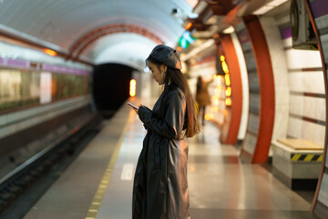 Korean girl with smartphone at empty metro platform. Stylish asian female browsing social media waiting for underground train. Young woman dependent from mobile phone and online communication concept