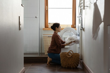 Full length tidy young indian ethnic woman folding white laundry clothes bedside towels in modern washing machine, doing household activities in light bathroom, domestic cleaning service concept.