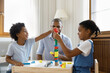 © fizkes - Happy Black kids playing together building high plastic tower on desk for board game, completing model from toy construction blocks. Dad watching little sibling children at home. Childhood, daycare
