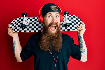 Canvas Print - Redhead man with long beard holding skate wearing headphones angry and mad screaming frustrated and furious, shouting with anger looking up.