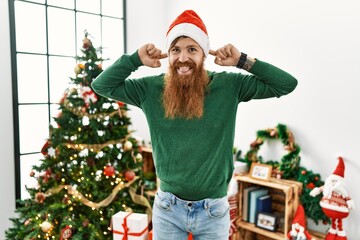 Wall Mural - Redhead man with long beard wearing christmas hat by christmas tree smiling pulling ears with fingers, funny gesture. audition problem