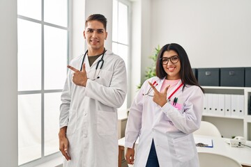 Poster - Young doctors wearing uniform and stethoscope at the clinic smiling cheerful pointing with hand and finger up to the side