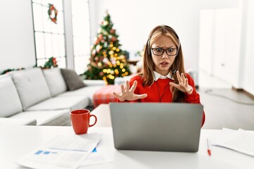 Poster - Young caucasian girl sitting on the table working using laptop by christmas tree afraid and terrified with fear expression stop gesture with hands, shouting in shock. panic concept.