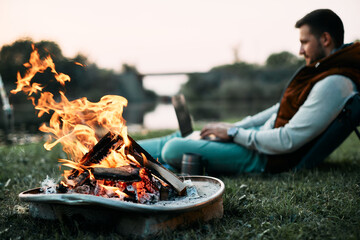 Wall Mural - close-up of campfire with man using laptop in the background.