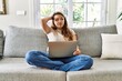 Beautiful young brunette woman sitting on the sofa using computer laptop at home confuse and wonder about question. uncertain with doubt, thinking with hand on head. pensive concept.
