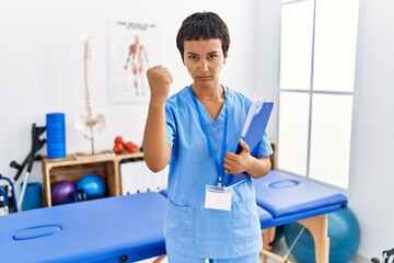 Poster - Young hispanic woman with short hair working at pain recovery clinic angry and mad raising fist frustrated and furious while shouting with anger. rage and aggressive concept.