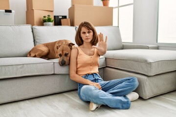 Poster - Young caucasian girl wearing casual clothes sitting on the floor with dog at home with open hand doing stop sign with serious and confident expression, defense gesture