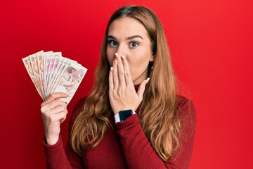 Poster - Young blonde woman holding turkish lira banknotes covering mouth with hand, shocked and afraid for mistake. surprised expression