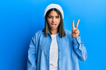 Poster - Young latin woman wearing cute wool cap smiling with happy face winking at the camera doing victory sign with fingers. number two.