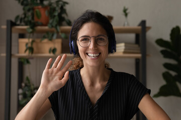 Wall Mural - Headshot screen portrait of friendly young latin lady in glasses headset teacher blogger look at digital camera wave hand. Millennial female coach in earphones talk to webcam starting distant training