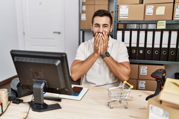 Wall Mural - Handsome hispanic man working at small business commerce laughing and embarrassed giggle covering mouth with hands, gossip and scandal concept