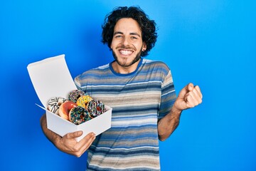 Sticker - Handsome hispanic man holding tasty colorful doughnuts screaming proud, celebrating victory and success very excited with raised arm