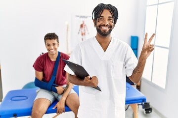 Canvas Print - Young hispanic man working at pain recovery clinic with a man with broken arm smiling with happy face winking at the camera doing victory sign with fingers. number two.