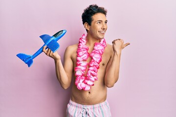 Young handsome man wearing swimsuit and hawaiian lei holding airplane toy pointing thumb up to the side smiling happy with open mouth