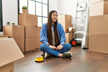 Wall Mural - Young chinese girl sitting on the floor at new home looking away to side with smile on face, natural expression. laughing confident.