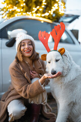 Wall Mural - Woman playing with her cute white dog near car and illuminated Christmas tree on a rooftop on nature at dusk. Concept of celebrating New Year holidays. Idea of Christmas mood and fun