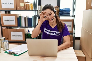 Poster - Young brunette woman wearing volunteer t shirt working at call center smiling happy doing ok sign with hand on eye looking through fingers