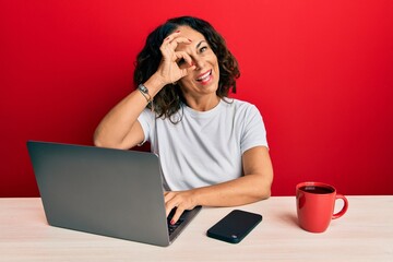 Sticker - Beautiful middle age woman working at the office using computer laptop smiling happy doing ok sign with hand on eye looking through fingers