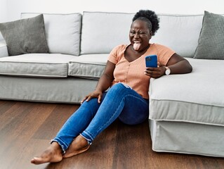 Canvas Print - Young african woman using smartphone sitting on the floor at home sticking tongue out happy with funny expression. emotion concept.